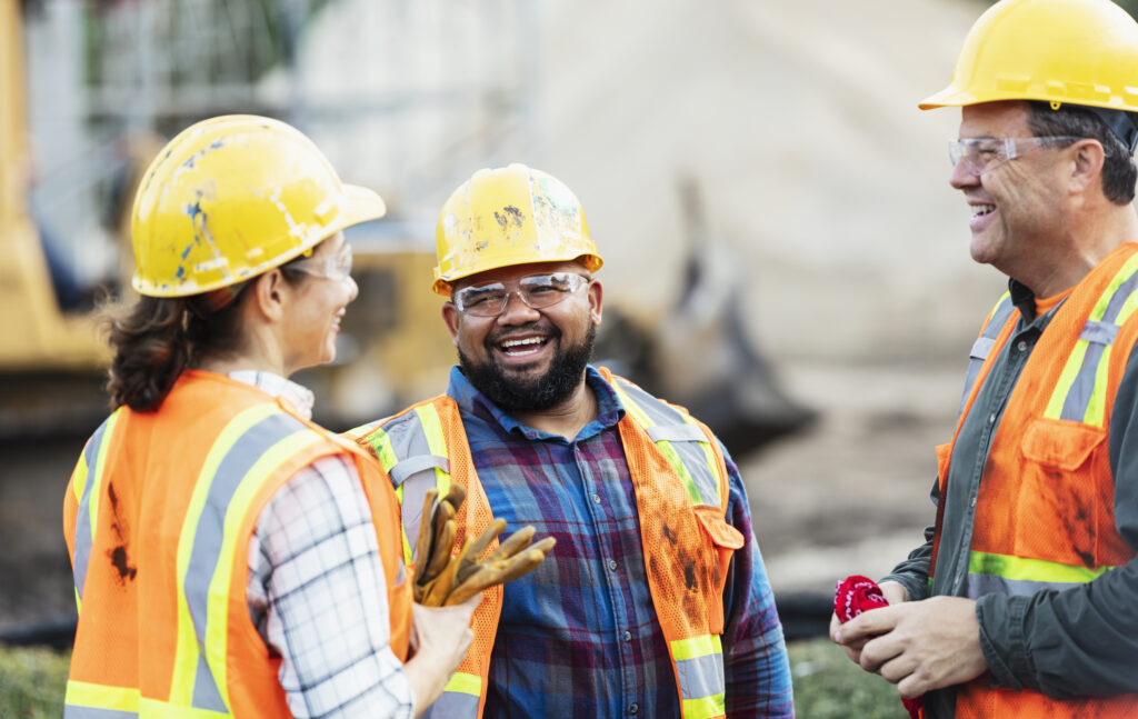 A group of three multi-ethnic workers at a construction site wearing hard hats, safety glasses and reflective clothing, smiling and conversing. The main focus is on the mixed race African-American and Pacific Islander man in the middle. The other two construction workers, including the woman, are Hispanic.