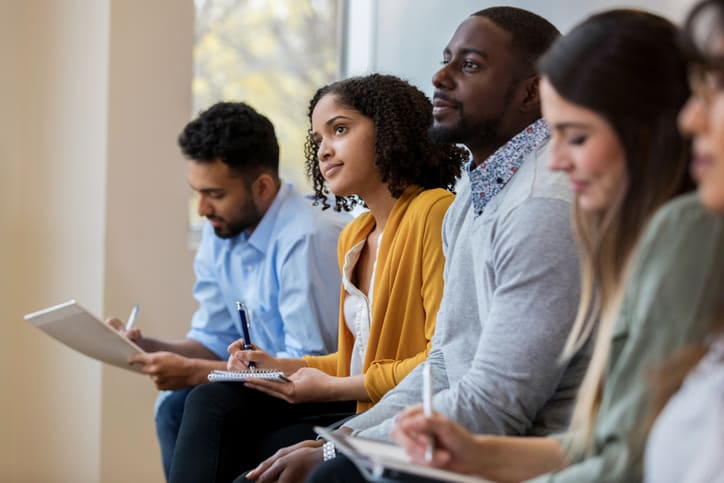 A group of business people sit in a row in a training class. They look at an unseen speaker as they concentrate on his lecture.